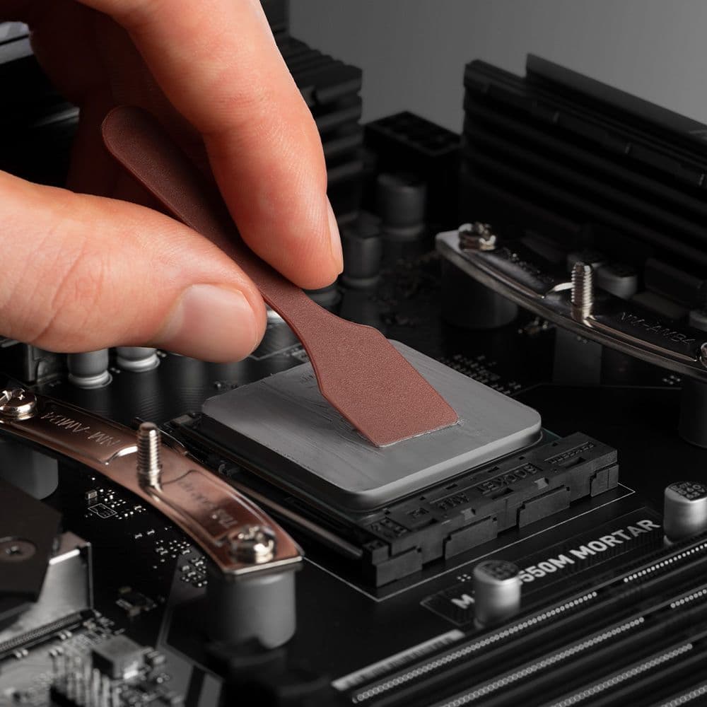 A hand using a brown Noctua spatula to spread a thin layer of thermal paste evenly over the surface of a CPU.